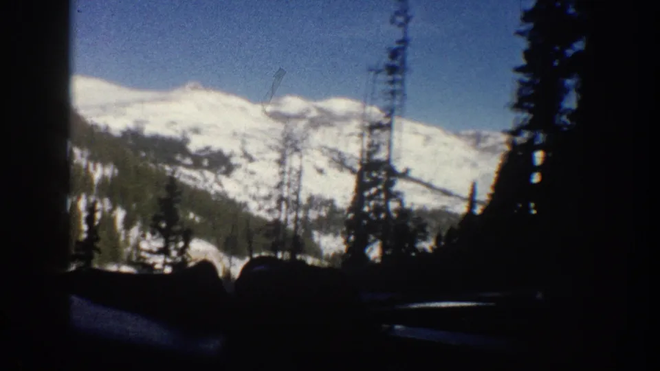 8mm home movie frame from Aspen, Colorado, 1961, showing a broad snow-covered summit dominating the view through a car window with dark conifer trees framing the lower portion.