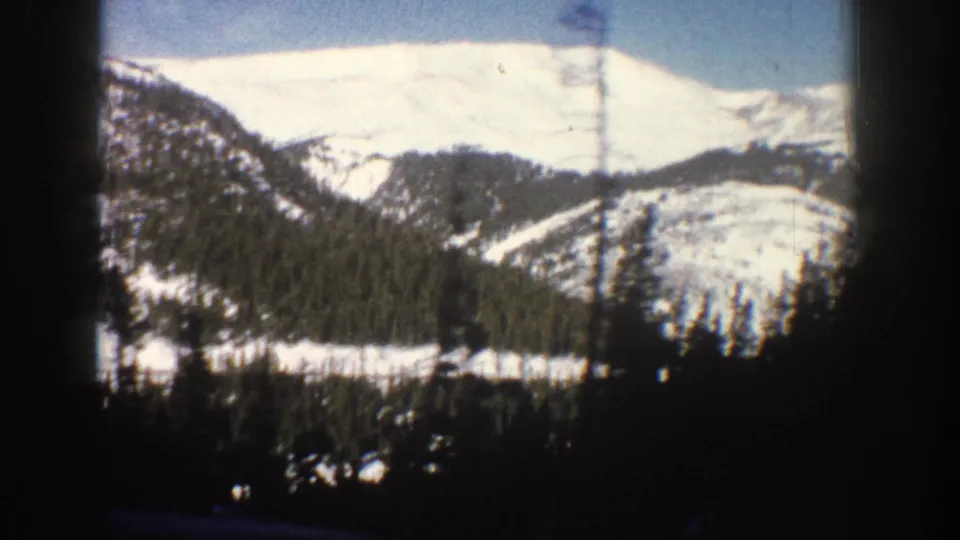 8mm home movie frame from Aspen, Colorado, 1961, showing a broad snow-covered mountain face with a conifer treeline at its base visible through the car window, with a dark film border at the right edge.