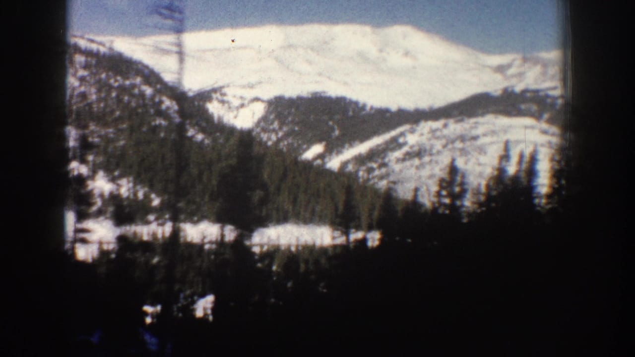 8mm home movie frame from Aspen, Colorado, circa 1961, showing a snow-covered Rocky Mountain peak with evergreen slopes and a dark window-edge element in the foreground.