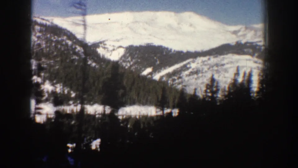 8mm home movie frame from Aspen, Colorado, circa 1961, showing a snow-covered Rocky Mountain peak with evergreen slopes and a dark window-edge element in the foreground.