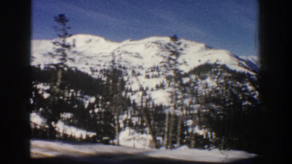 8mm home movie frame circa 1961 showing a wide Rocky Mountain winter panorama near Aspen, Colorado, with snow-covered peaks, conifer forest, and clear blue sky.