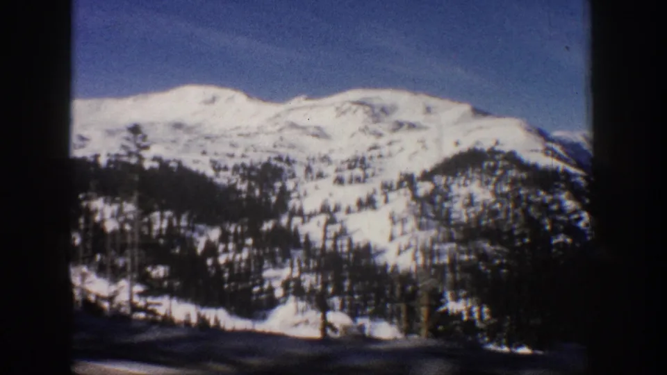 8mm home movie frame circa 1961 showing a wide Colorado Rocky Mountain winter scene with deep blue sky, snow-covered peaks, and a single pine silhouette at lower left.