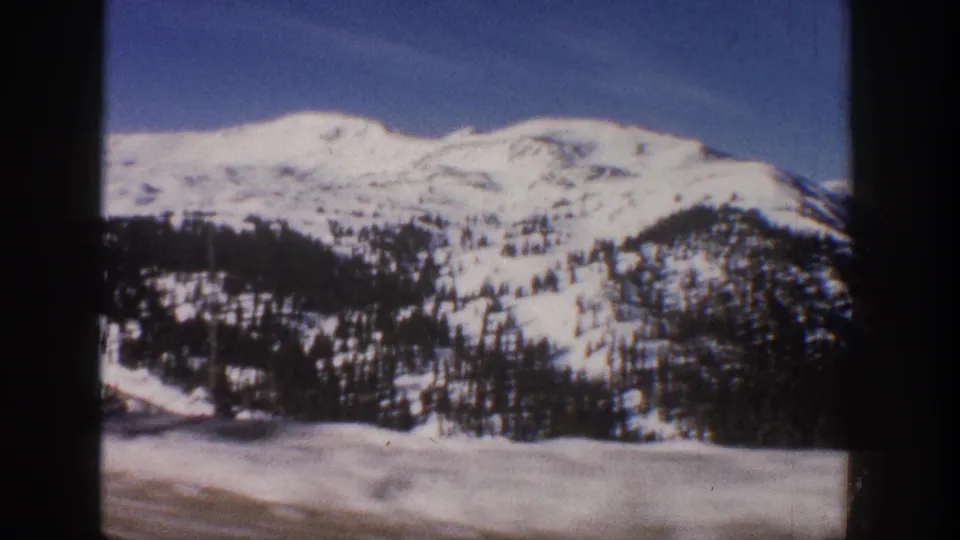 8mm home movie frame circa 1961 showing the same Colorado Rocky Mountain winter panorama with snow-covered peaks, evergreen forest, and open snowy valley near Aspen.