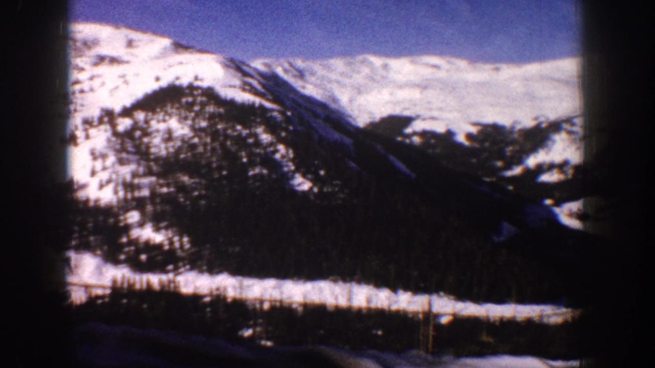 Continuing pan of snow-covered mountain ridge with forested lower slopes under blue sky, 8mm film-gate borders visible.
