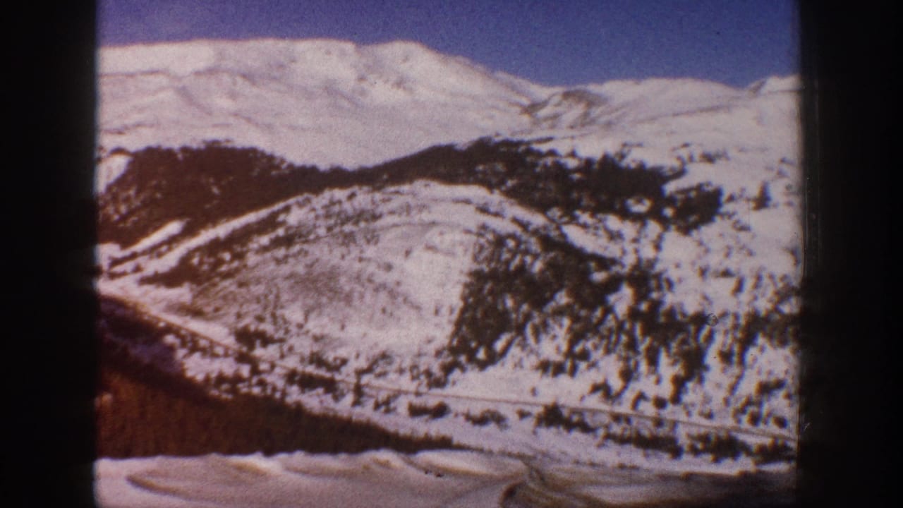 Continued pan across snow-laden mountain slopes with exposed rocky faces and a winding road curving through the valley.