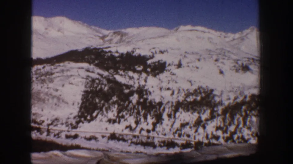 8mm wide shot of snow-covered hillside with dense conifer forest and clear blue sky, Aspen, Colorado area, circa 1961.
