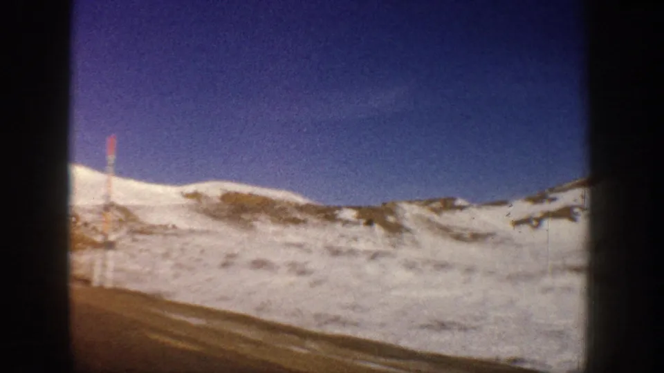Mountain road running through snow-covered slopes with a red roadside marker stake, deep blue sky, Colorado Rockies, circa 1961, 8mm.