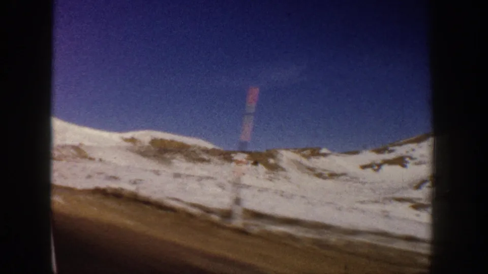 Snowy mountain road with red roadside marker stake and blue-purple emulsion color shift, filmed from a moving vehicle, Colorado Rockies area, circa 1961.