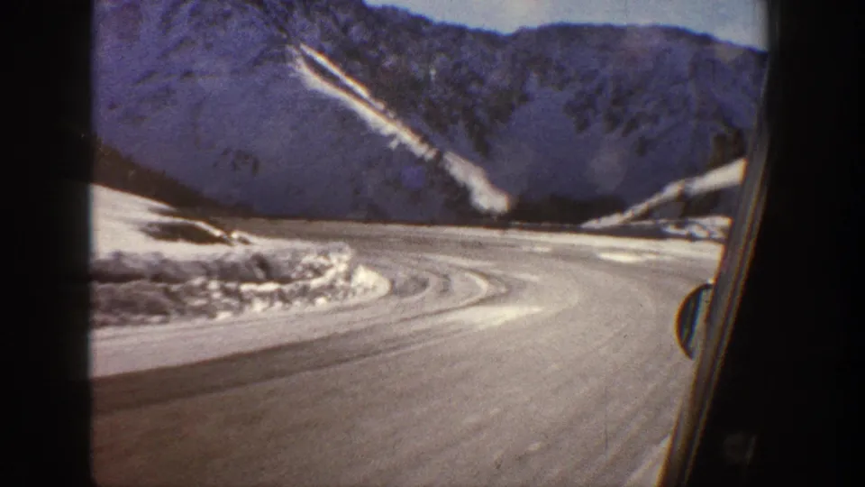 Hairpin curve on a snowy mountain road with rocky cliff faces, filmed through a car window, Colorado Rockies, circa 1961, 8mm.