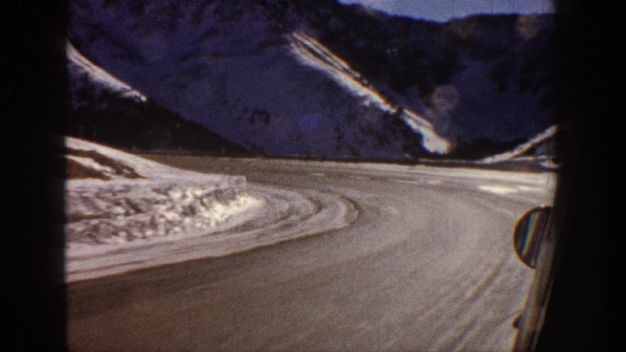 Winding mountain road curving right through snow-covered terrain, partial car panel visible at lower frame edge.