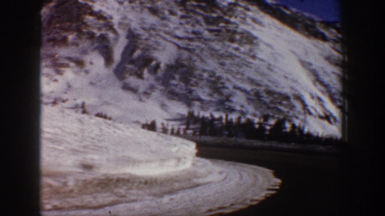 8mm home movie frame from 1961 showing a winding road through heavy snowbanks with a snow-covered Rocky Mountain peak in the background, Aspen area, Colorado.