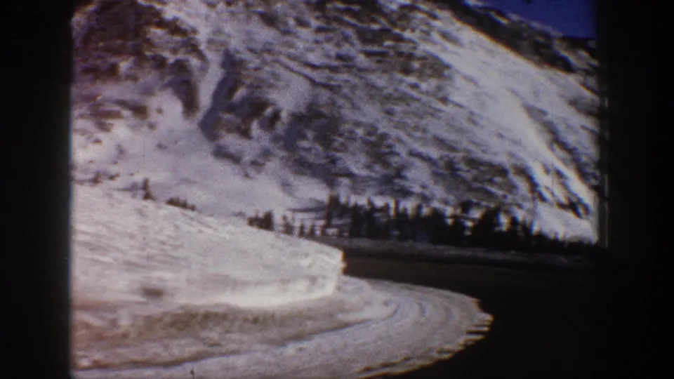 8mm home movie frame from 1961 showing a winding road through heavy snowbanks with a snow-covered Rocky Mountain peak in the background, Aspen area, Colorado.