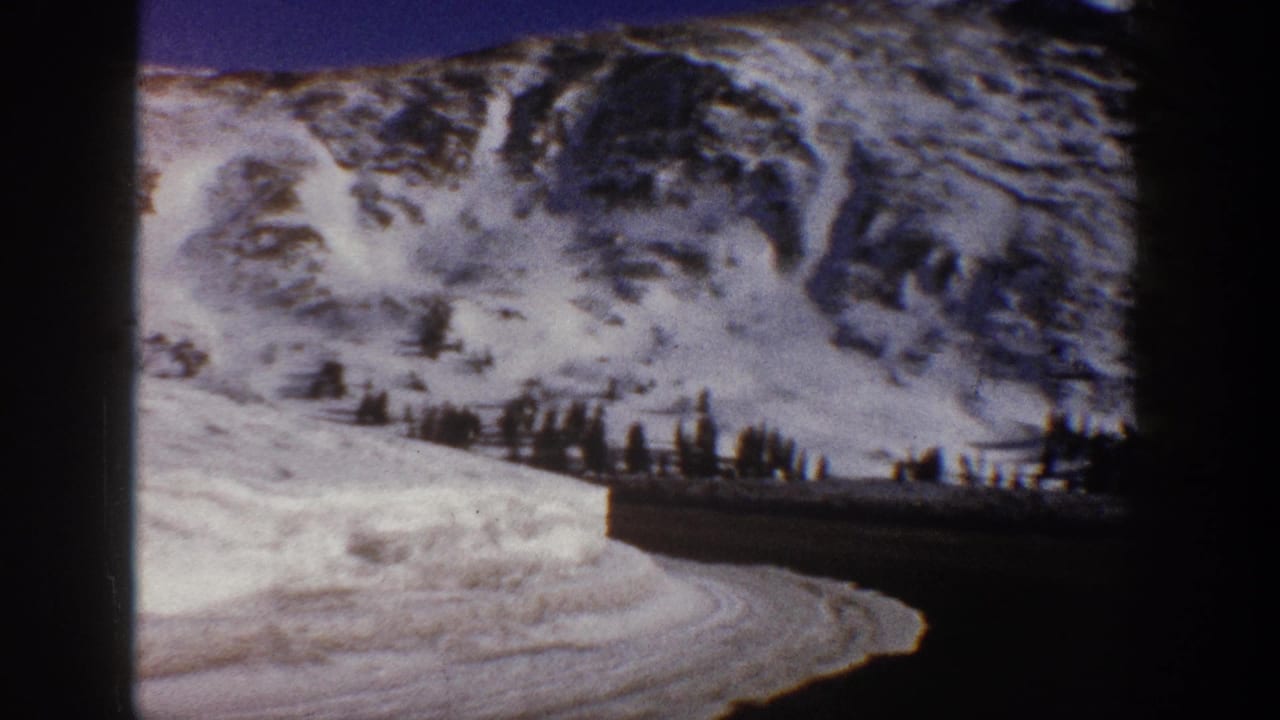 Steep snow-covered mountain peak fills the upper frame as the road curves below, with an evergreen treeline at the mountain base.