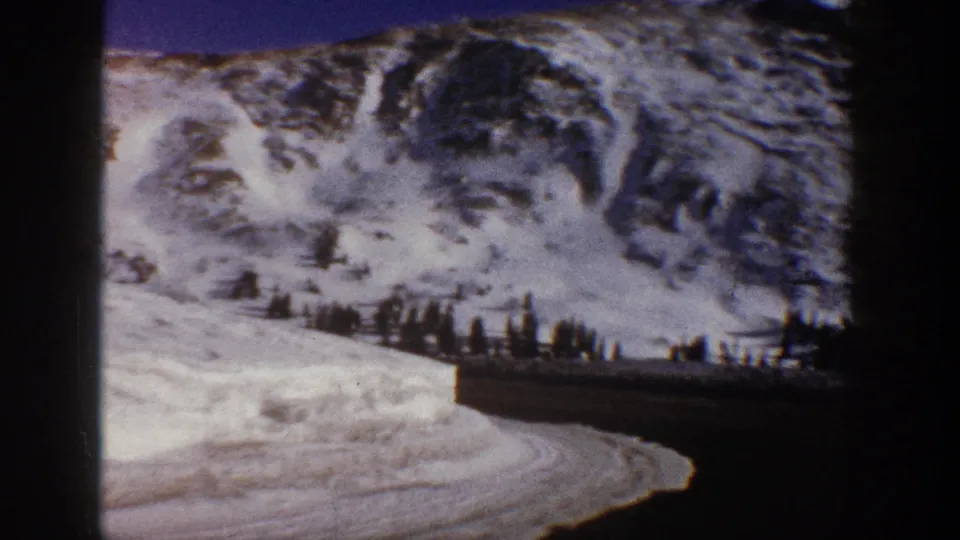 8mm frame from 1961 showing a steep snow-covered peak and curving mountain road below a deep blue sky, near Aspen, Colorado, as noted in clip metadata.