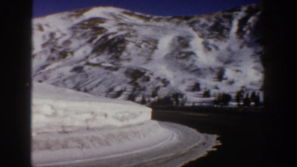 8mm home movie frame from 1961 showing a snow-patched mountain hillside with warm earthy tones and a curving road in the foreground, Aspen area, Colorado.