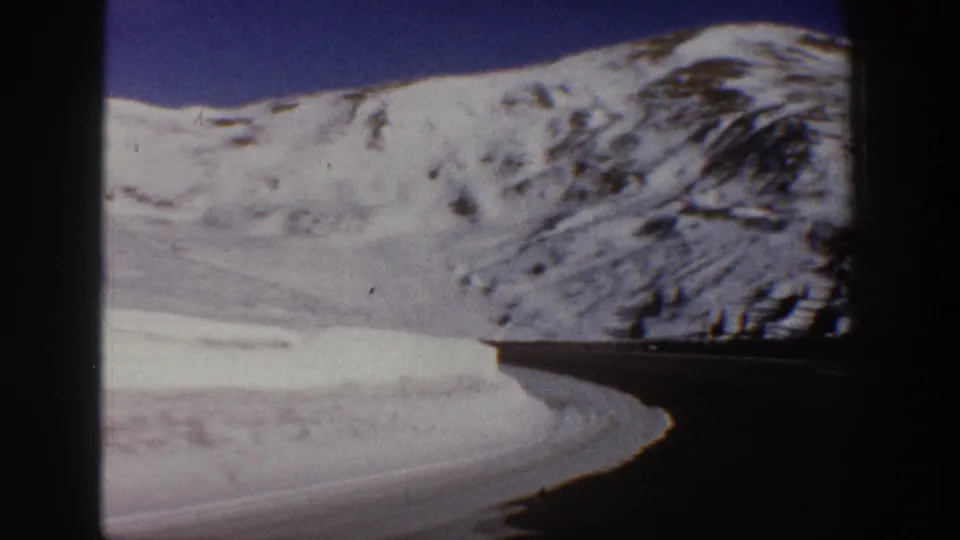 8mm frame from 1961 capturing a wide snowy valley near Aspen, Colorado, with a mountain peak at right, deep blue sky, and a curving road along the lower foreground.