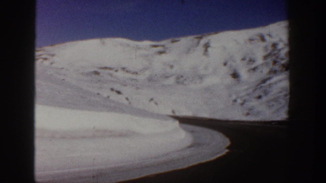 8mm home movie frame, c.1961, showing a winding road curving through snow-covered Colorado mountain terrain with a deep blue sky above.