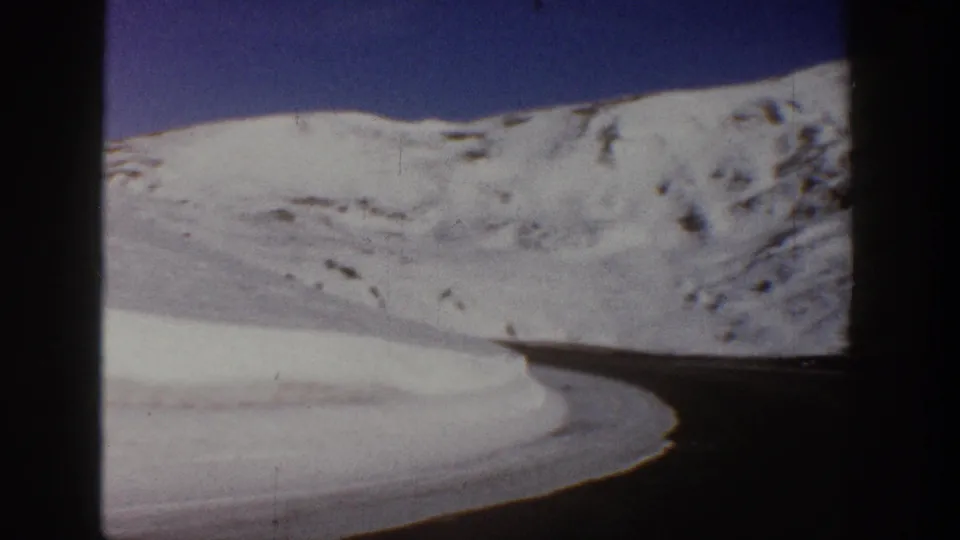 8mm home movie frame, c.1961, showing a winding road curving through snow-covered Colorado mountain terrain with a deep blue sky above.