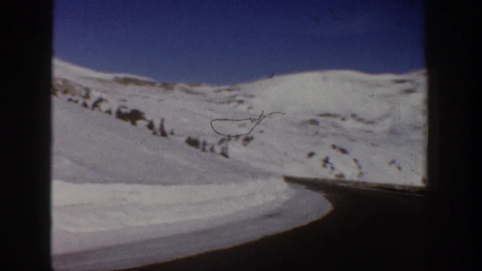 8mm driving POV c.1961, looking ahead along a curved mountain road banked by deep snow, with bare branches at upper right and blue sky, consistent with the Aspen, Colorado area.