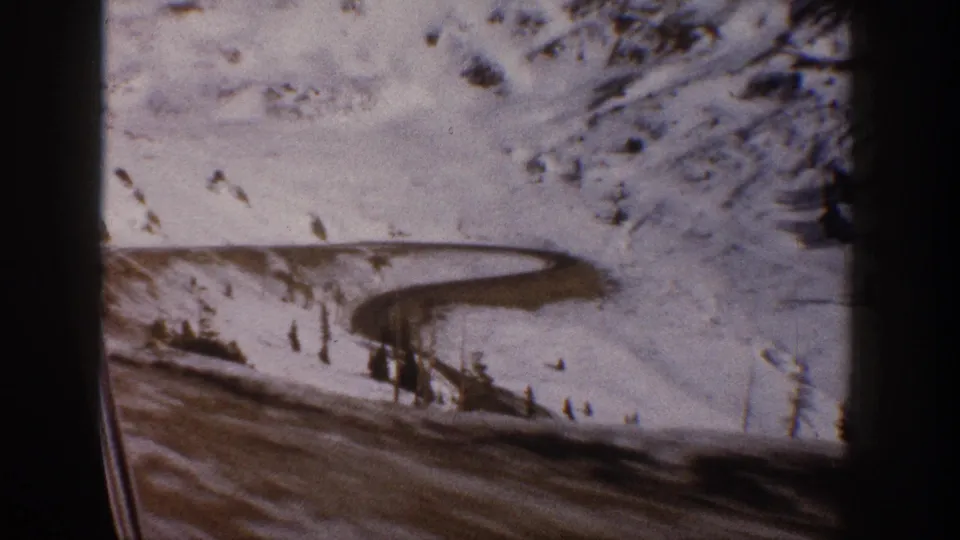 8mm frame c.1961 showing a winding mountain road through snow with sparse conifers at right; dark vehicle foreground suggests a moving driving shot, Aspen area, Colorado.