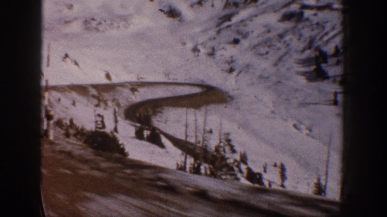 8mm film frame circa 1961 showing a winding road through a snowy Colorado mountain valley with scattered conifers, warm amber tones, and visible film grain.