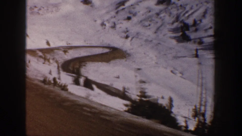 8mm film frame circa 1961 showing a winding road through a snow-covered Colorado mountain valley with evergreen trees, bare slope patches, and amber film tones.