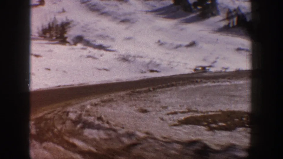 8mm film frame circa 1961 showing a snow-edged mountain road curving through a winter landscape in the Aspen, Colorado area with conifers above.