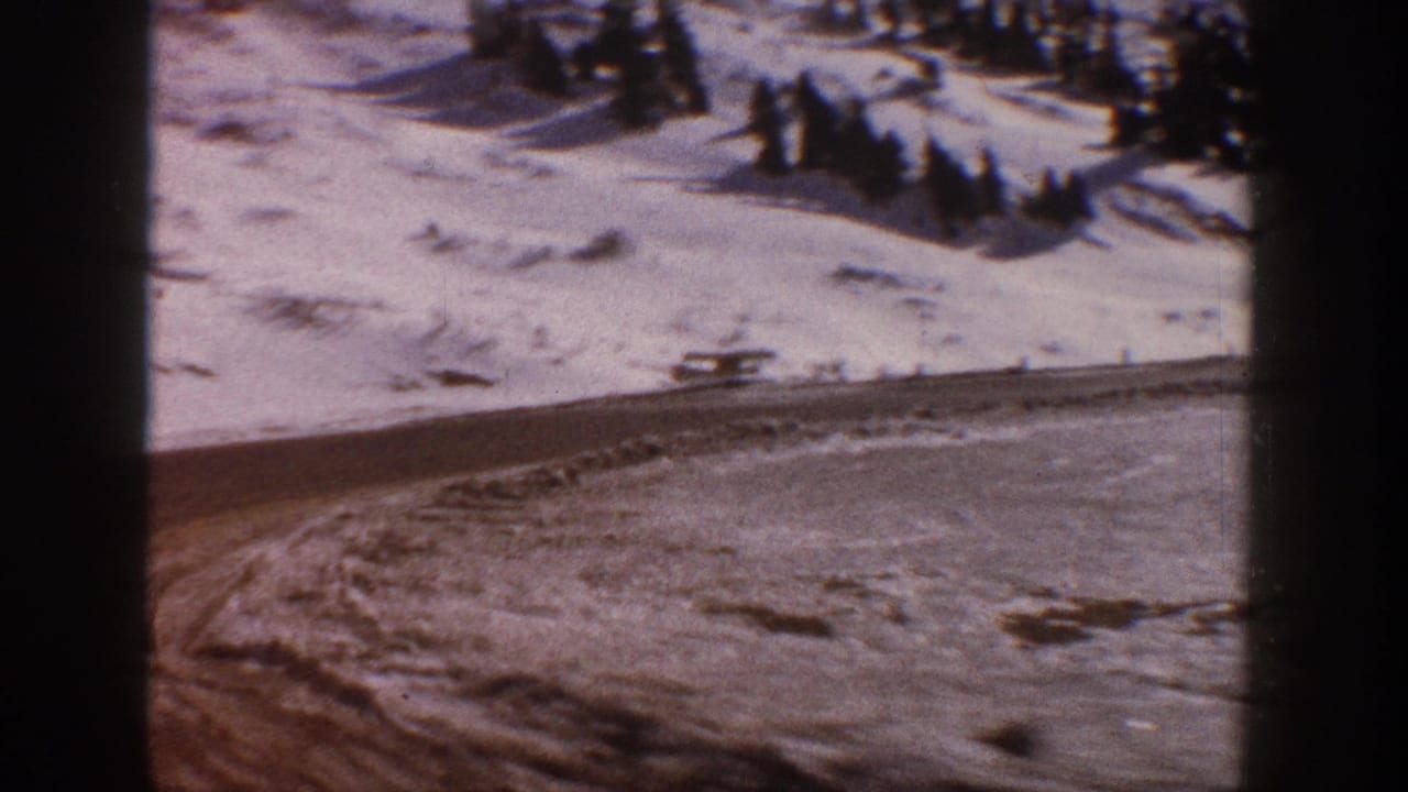 8mm home movie frame, circa 1961, showing a snow-covered mountain slope near Aspen, Colorado, with a winding road and dark foreground visible from a passing vehicle.