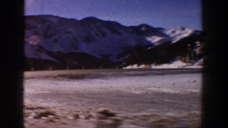 8mm home movie frame, circa 1961, showing a broad snow-covered mountain range under vivid blue sky near Aspen, Colorado, with a frozen valley floor in the foreground.