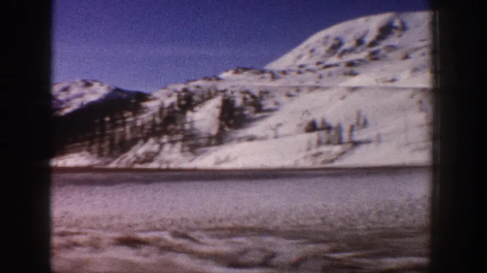8mm home movie frame, circa 1961, showing snow-laden mountain slopes with evergreen trees near Aspen, Colorado, a frozen lake in the foreground, and clear blue sky above.
