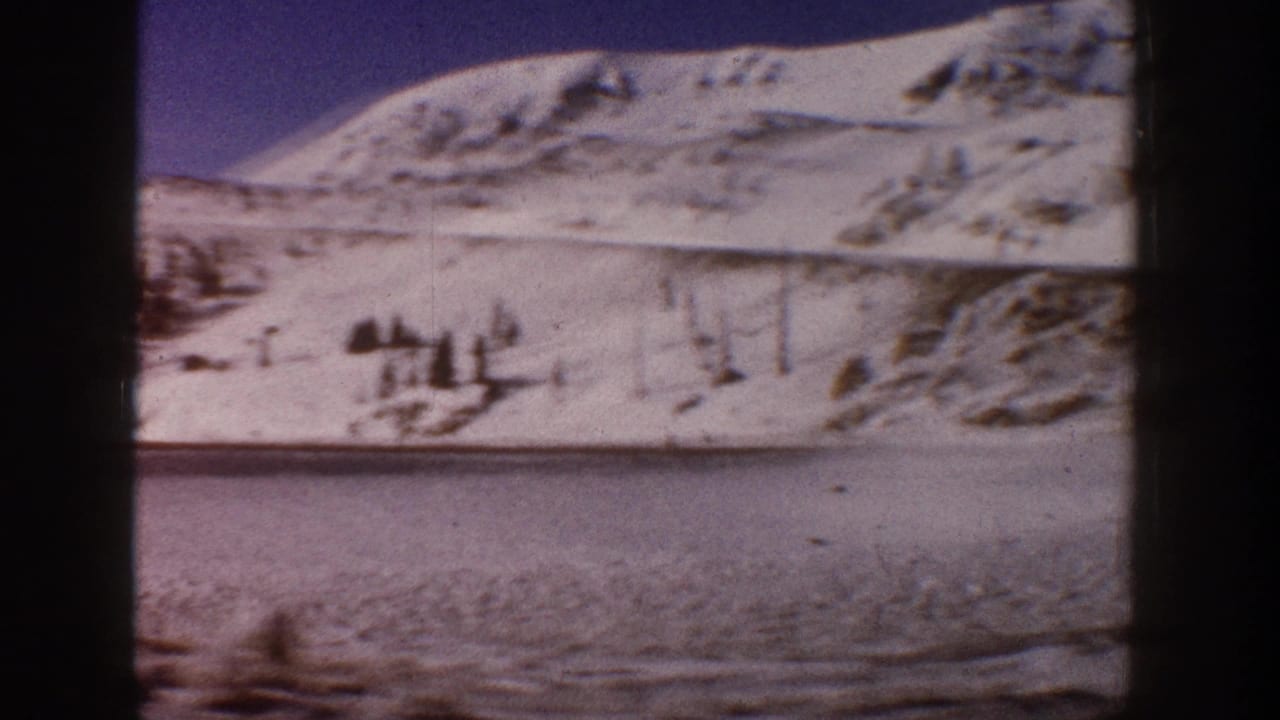1961 Aspen, Colorado: snow-covered mountain ridgeline above a flat frozen or snow-blanketed valley, shot on 8mm home movie film with dark edge vignette.