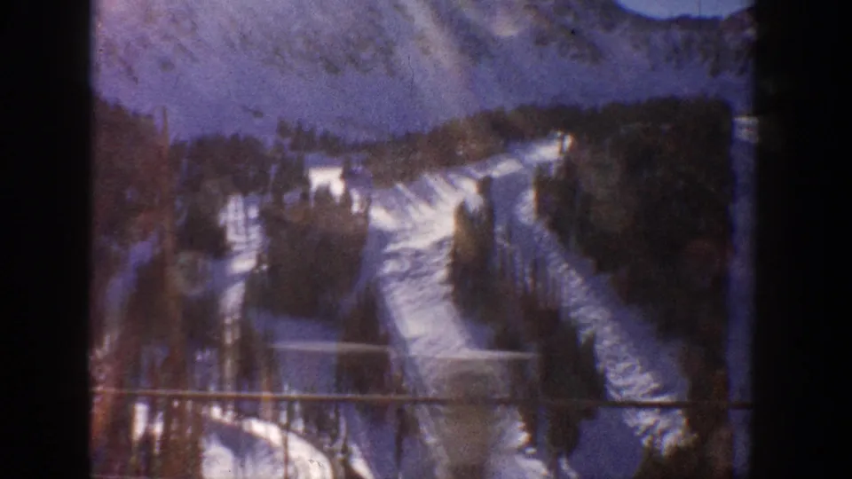 1961 Aspen, Colorado: elevated view down snow-covered ski runs flanked by dense conifer forest, shot on 8mm home movie film with visible grain and dark edge vignette.