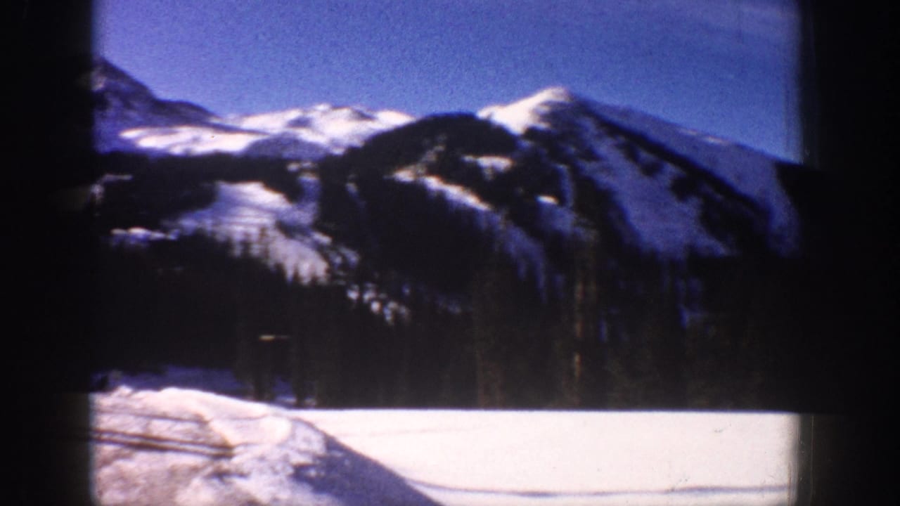 Snow-covered mountain range with evergreen forest and a snow-covered road or path visible below, filmed through a vehicle window.