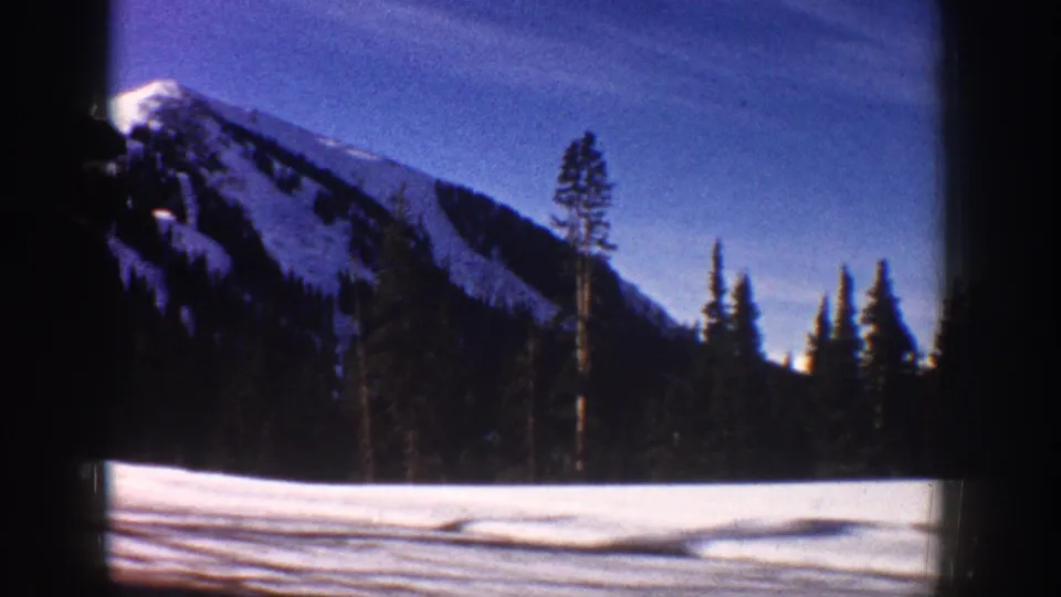 8mm footage of a prominent snow-covered mountain peak with tall silhouetted conifers in the foreground, viewed through a moving car window, Aspen area, circa 1961.