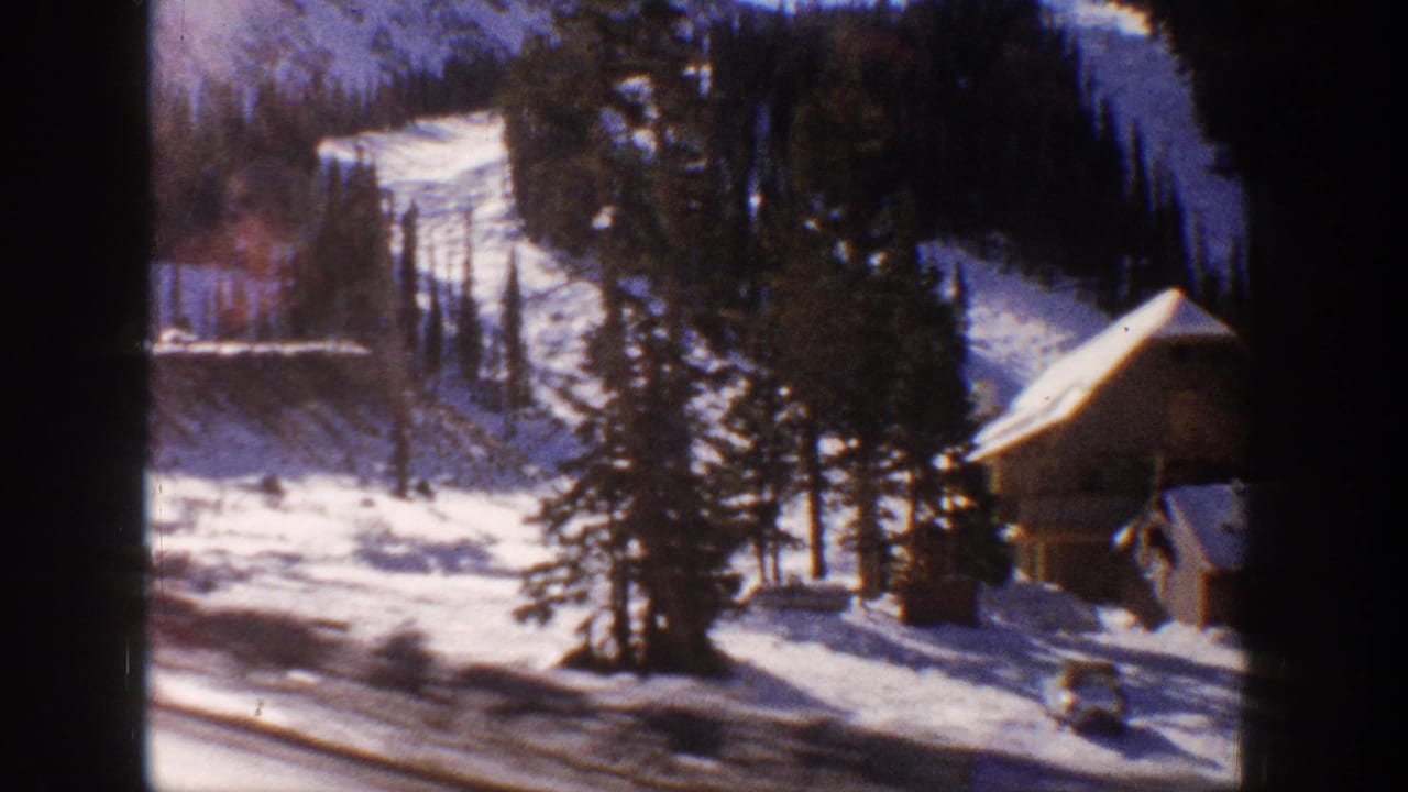 8mm home movie frame from Aspen, Colorado, 1961 — wooden chalet-style cabin amid snow-covered pines, vehicle parked on road below, forested mountain backdrop.