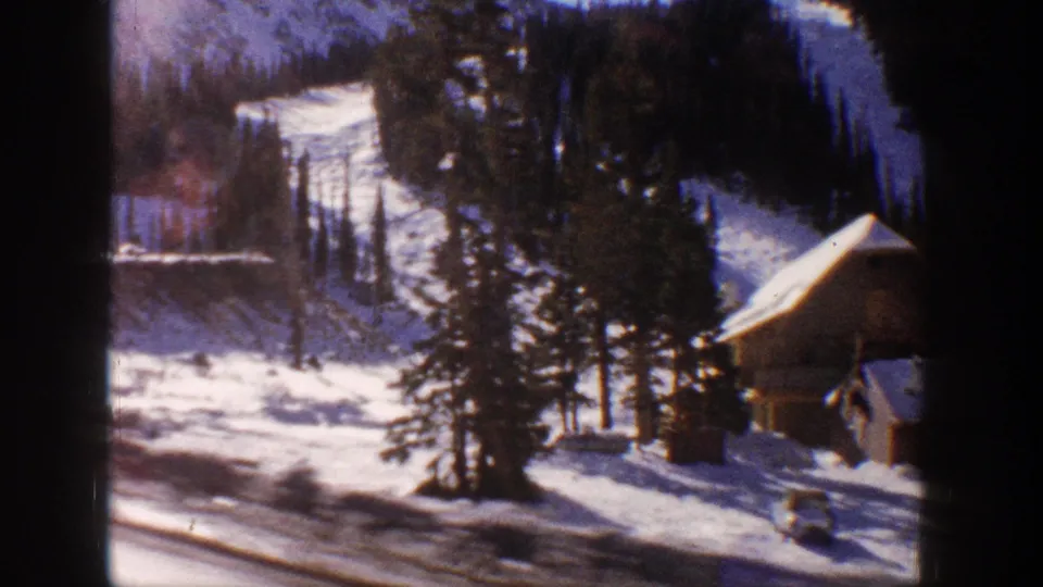 8mm home movie frame from Aspen, Colorado, 1961 — wooden chalet-style cabin amid snow-covered pines, vehicle parked on road below, forested mountain backdrop.