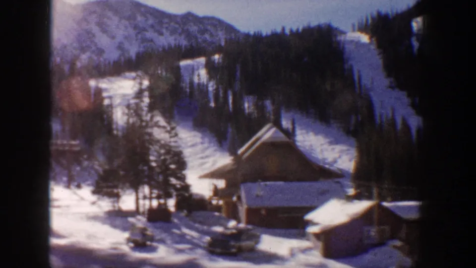 8mm frame showing lodge buildings and parked vehicles at what appears to be an Aspen ski resort, Colorado, 1961, with forested mountain slopes and open runs behind.