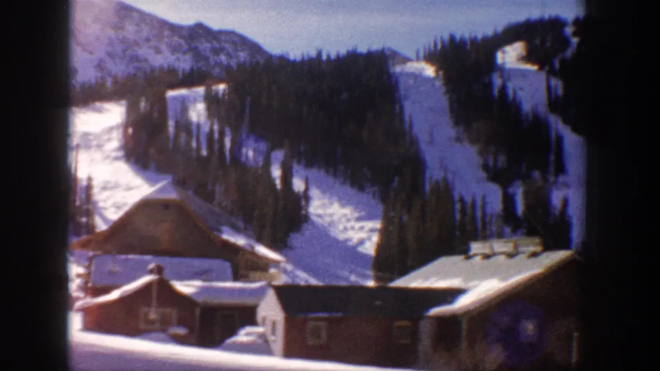 8mm home movie frame showing red-painted resort buildings below a large mountain with open ski slopes, Aspen area, Colorado, 1961; cooler blue-purple tones on snow.