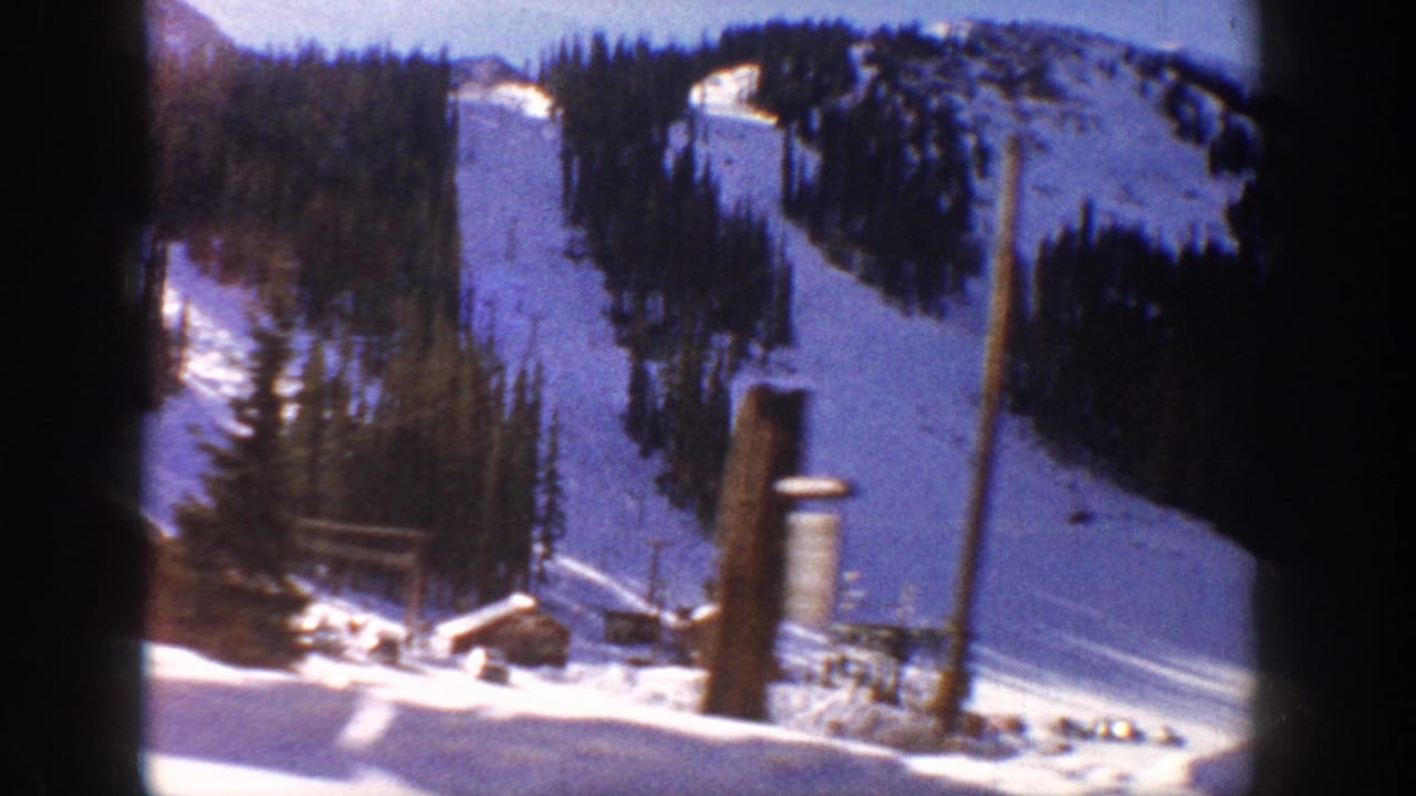 8mm home movie frame, possibly Aspen Colorado c.1961, showing wooden ski lift poles, a small base building, and snow-covered slopes lined with dense evergreen trees.