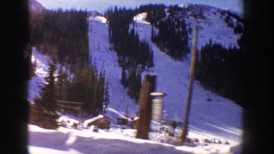 8mm home movie frame, possibly Aspen Colorado c.1961, showing wooden ski lift poles, a small base building, and snow-covered slopes lined with dense evergreen trees.