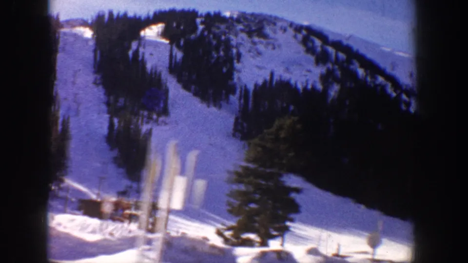 8mm home movie frame, possibly Aspen Colorado c.1961, showing two wooden ski lift towers, a base building at the slope foot, and forested mountain above under a pale sky.