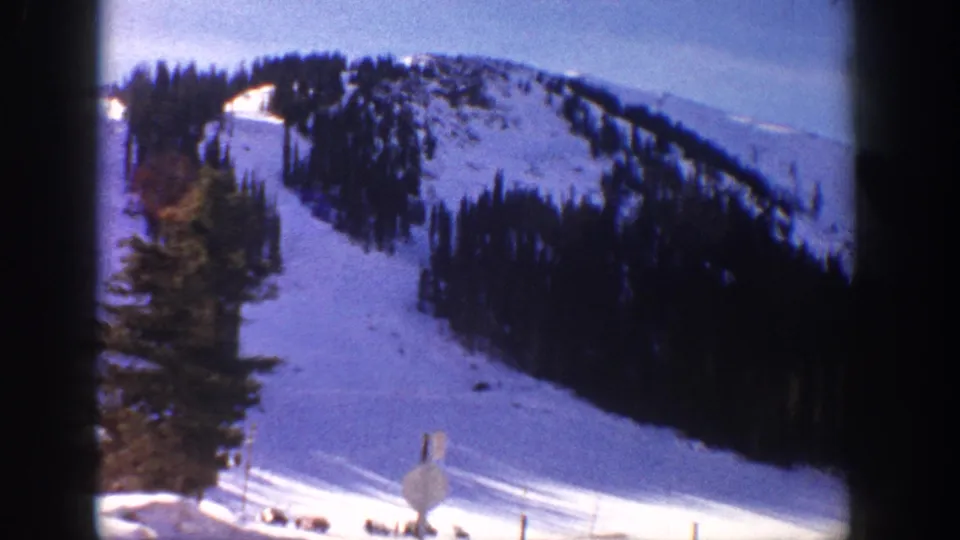 8mm home movie frame, possibly Aspen Colorado c.1961, showing a wide open ski run with a small signpost in the lower foreground, bordered by evergreen forest under blue sky.