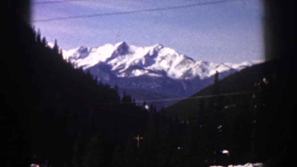 8mm home movie frame, possibly Aspen Colorado c.1961, showing a panoramic view of snow-capped mountain peaks in the distance, a dark forested ridge at right, and an expansive sky.