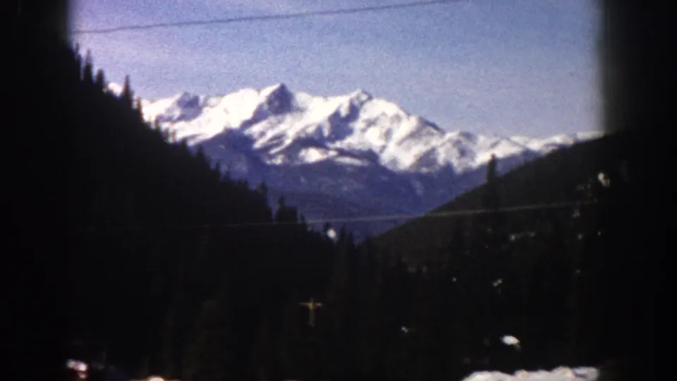 Rocky Mountain peaks above pine forest, road and vehicles visible at lower frame edge, 8mm driving footage, Aspen, Colorado area, circa 1961.