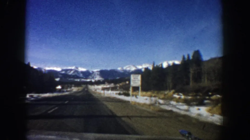 Forward driving POV on two-lane Colorado highway with illegible roadside sign, snow-dusted verges, and distant Rocky Mountain peaks, 8mm home movie, circa 1961.