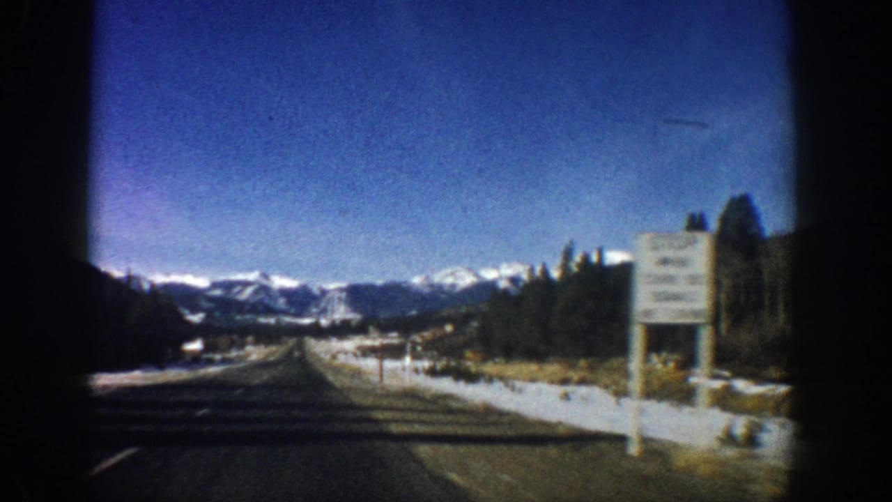 8mm home movie from a moving car on a Colorado highway, c.1961, with a roadside sign, snowy shoulders, evergreen trees, and Rocky Mountain backdrop.