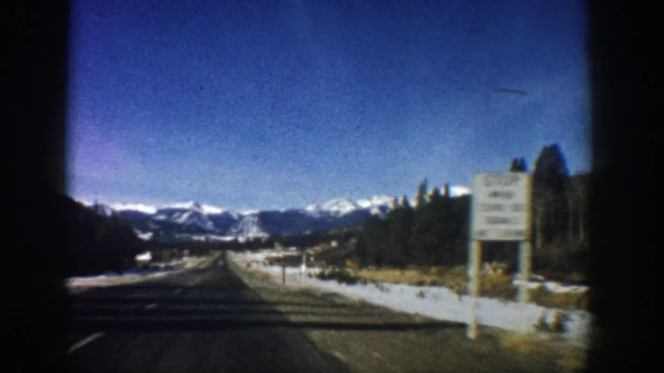 8mm home movie from a moving car on a Colorado highway, c.1961, with a roadside sign, snowy shoulders, evergreen trees, and Rocky Mountain backdrop.