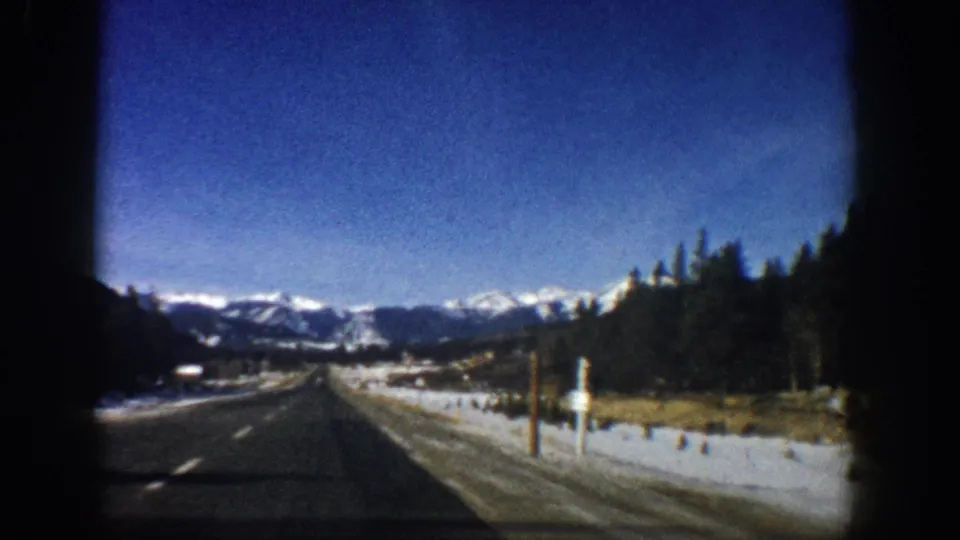 8mm home movie from moving vehicle on Colorado highway, c.1961, showing utility poles, snow-dusted shoulders, evergreen trees, and peaks under blue sky.