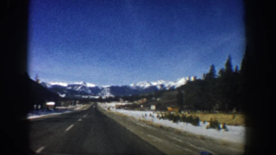 8mm film from moving vehicle, c.1961, Colorado Rockies, showing a curving two-lane highway with snowy shoulders, pine trees, and distant mountain peaks.