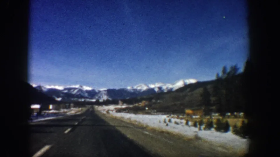 8mm home movie from moving car, c.1961, Colorado highway curving right with snowy shoulders, roadside vegetation, and snow-capped Rocky Mountains.